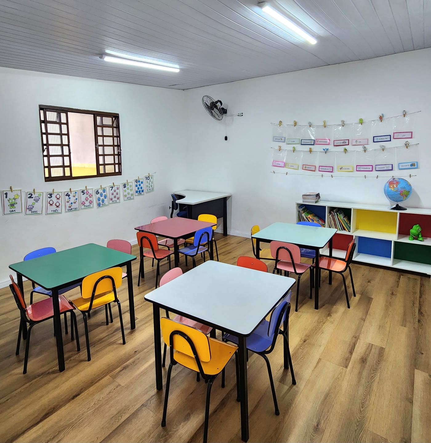 Espaço pedagógico com mesas coloridas, cadeiras infantis e estantes