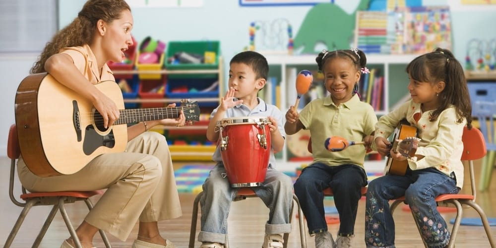 Educadora tocando violão e crianças com tambor e chocalhos em uma aula de música